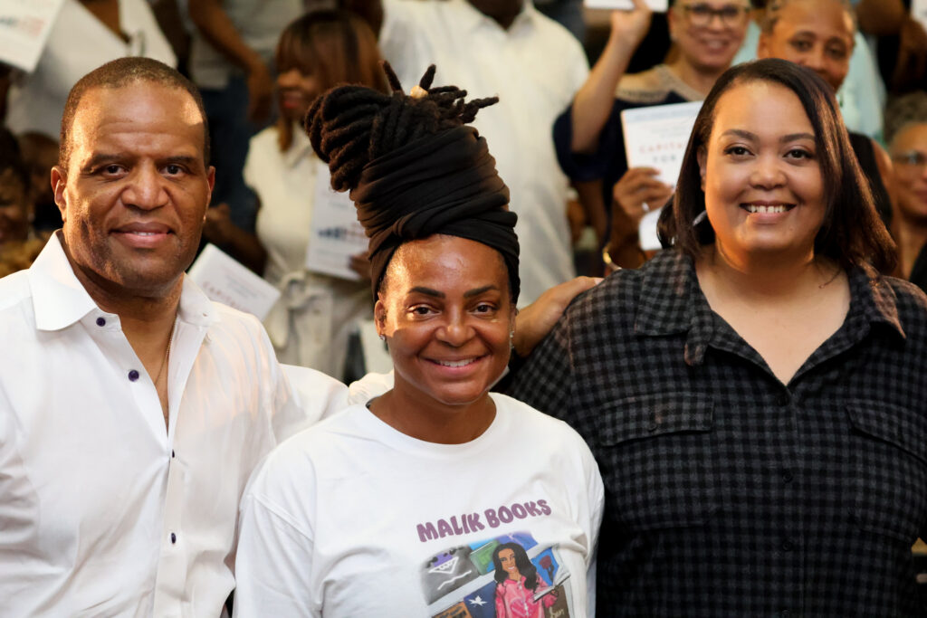 John Hope Bryant smiles with Malik Books supporters as the audience behind them holds up copies of Capitalism for All