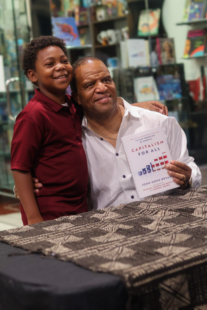 John Hope Bryant embraces a young boy while holding a copy of Capitalism for All