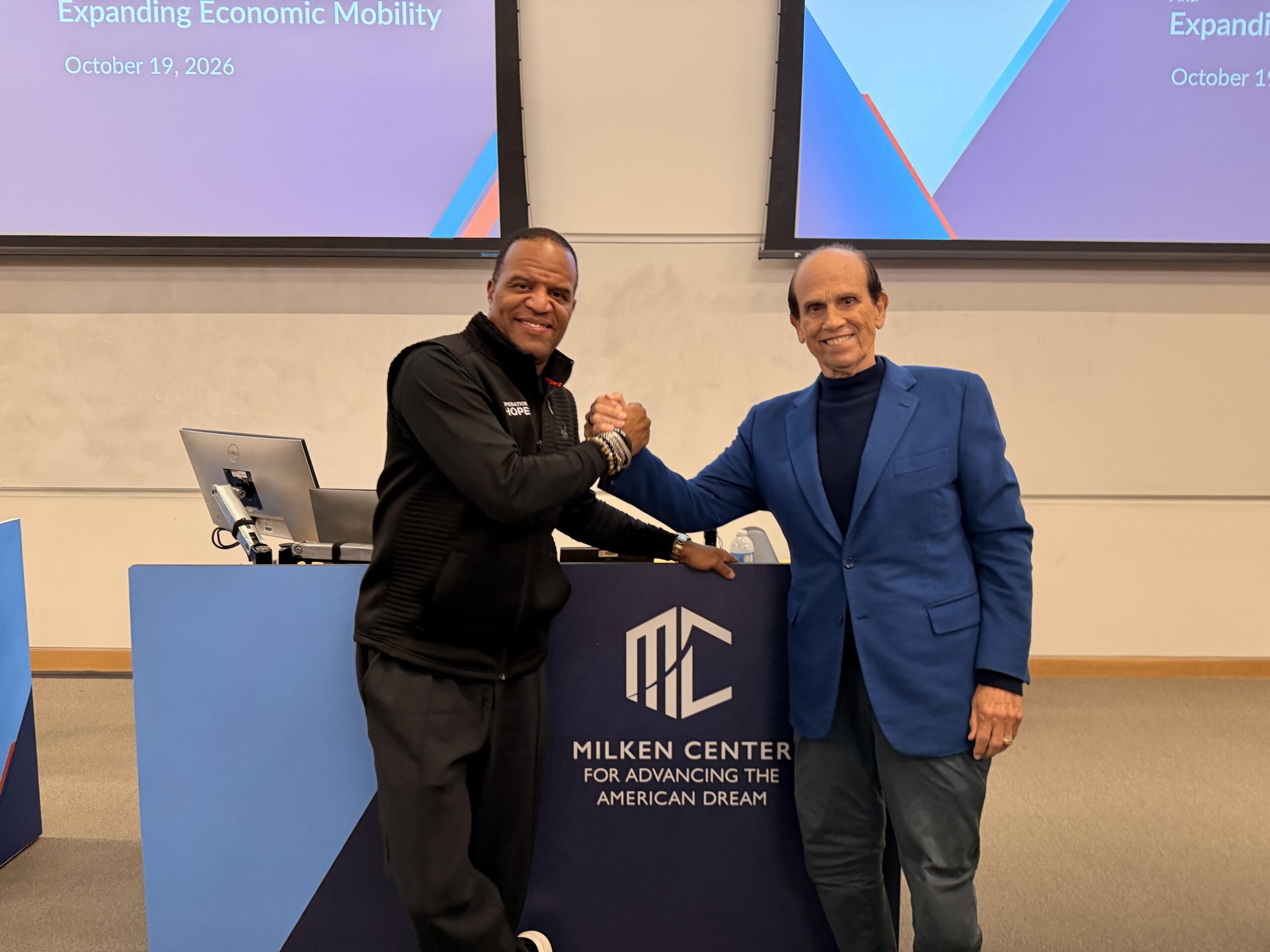 John Hope Bryant and Michael Milken shaking hands in front of the Milken Center for Advancing the American Dream backdrop at USC